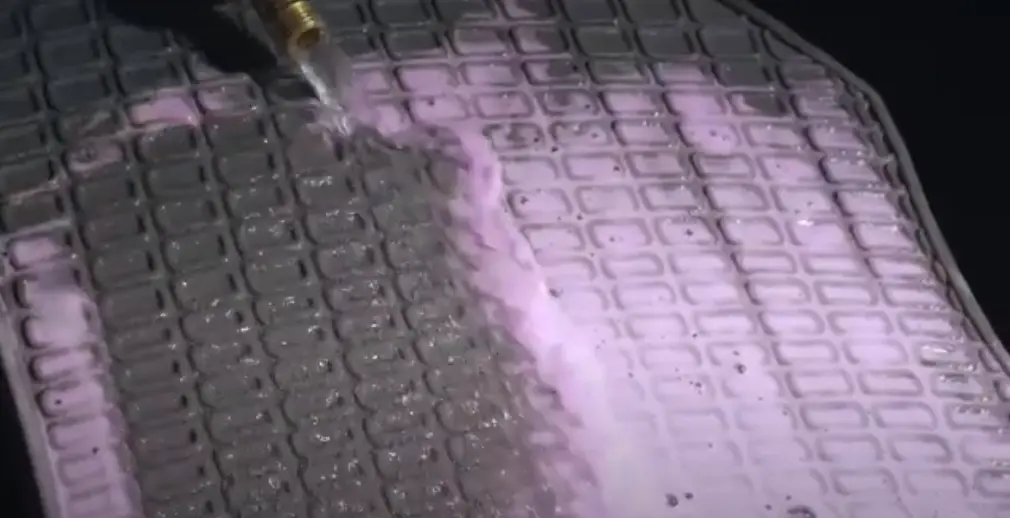 Pink liquid cleaner spread on a grey car floor mat and a hose pouring water on the mat